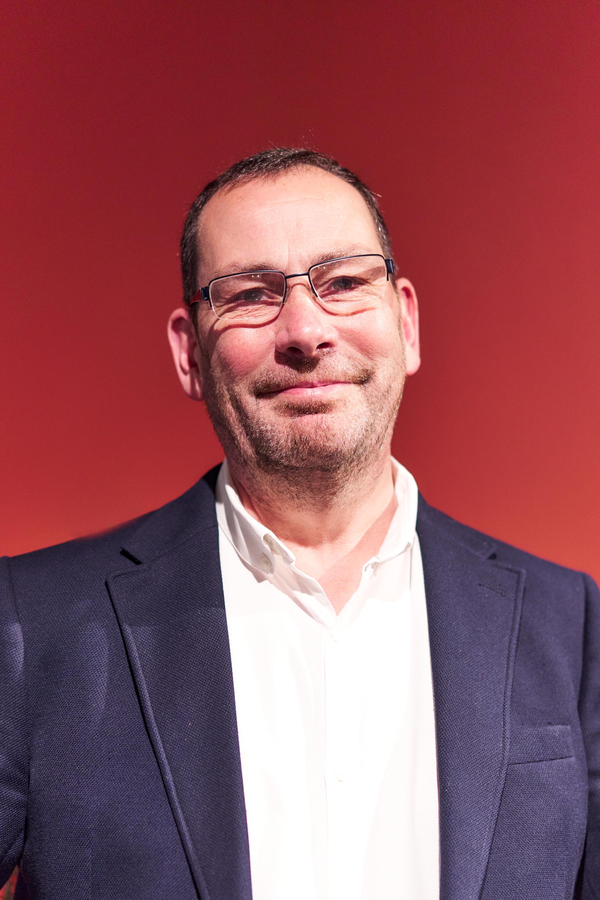 Man in a navy blazer and white shirt smiling, against a red background, at Material Source Studio Glasgow event.