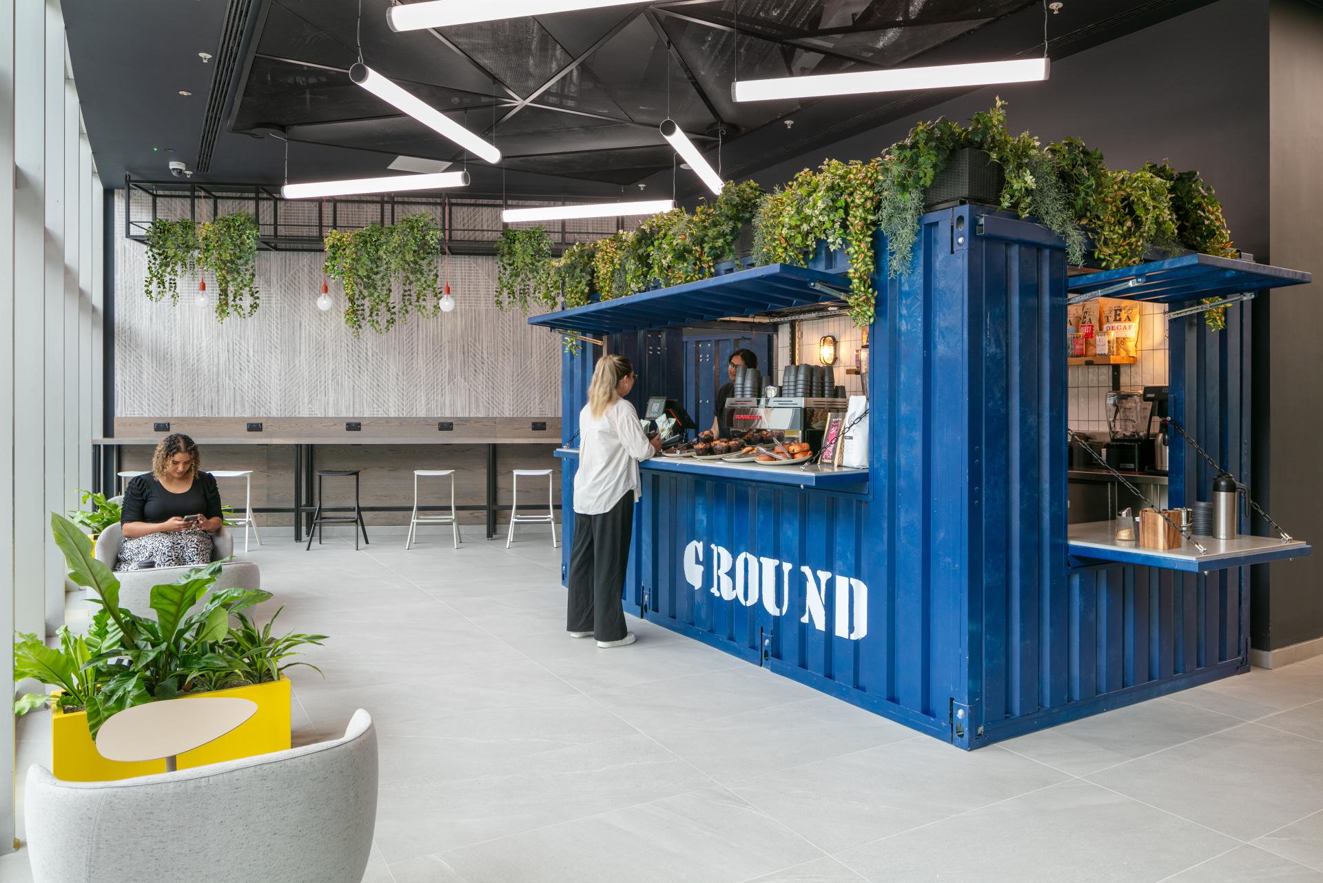 Modern office café featuring a vibrant blue shipping container serving food and drinks, emphasizing workplace culture and sustainability.
