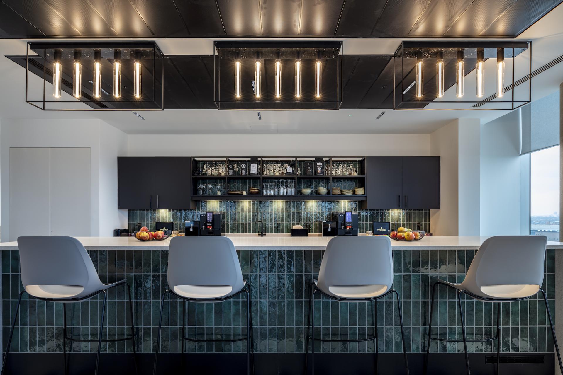 Modern office kitchen featuring a stylish bar area, green tiled backsplash, and contemporary lighting, reflecting workplace design trends.