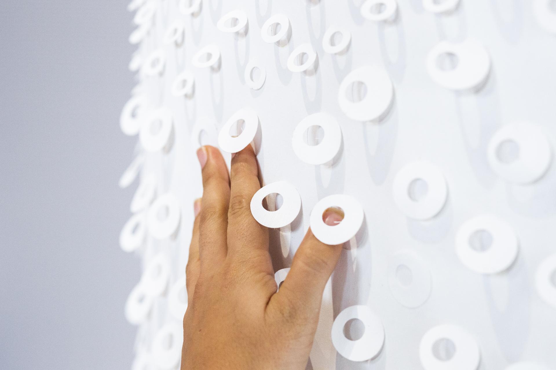 A hand gently touches an interactive, textured wall installation featuring layered white circular elements at a design exhibition.