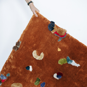 Soft, colorful Chaos Rug designed by Audronė Drungilaitė, showcasing abstract patterns in vibrant hues on a warm brown background.