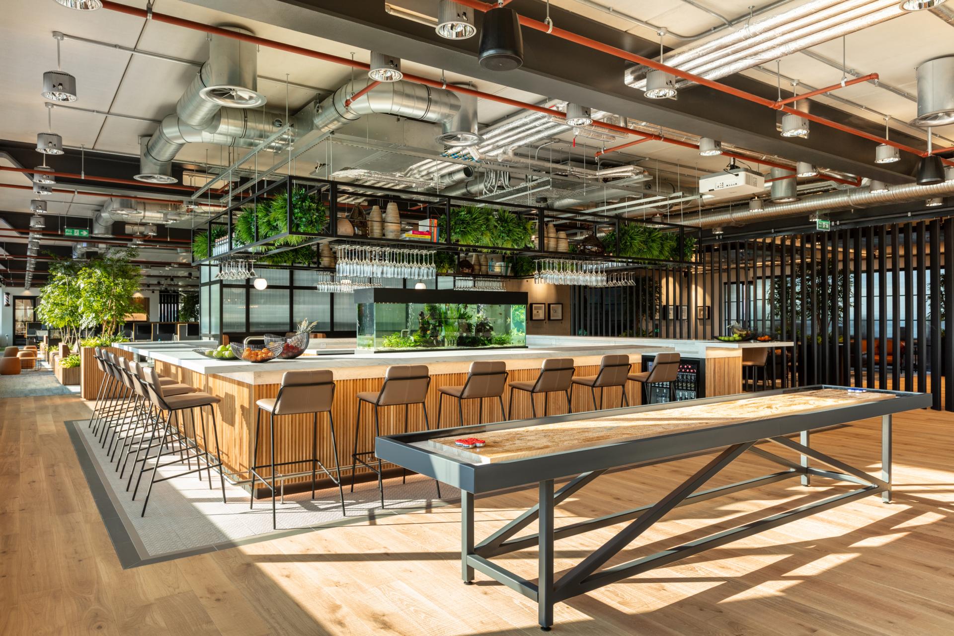 Modern office kitchen and casual dining area featuring greenery, bar seating, and a collaborative workspace design.