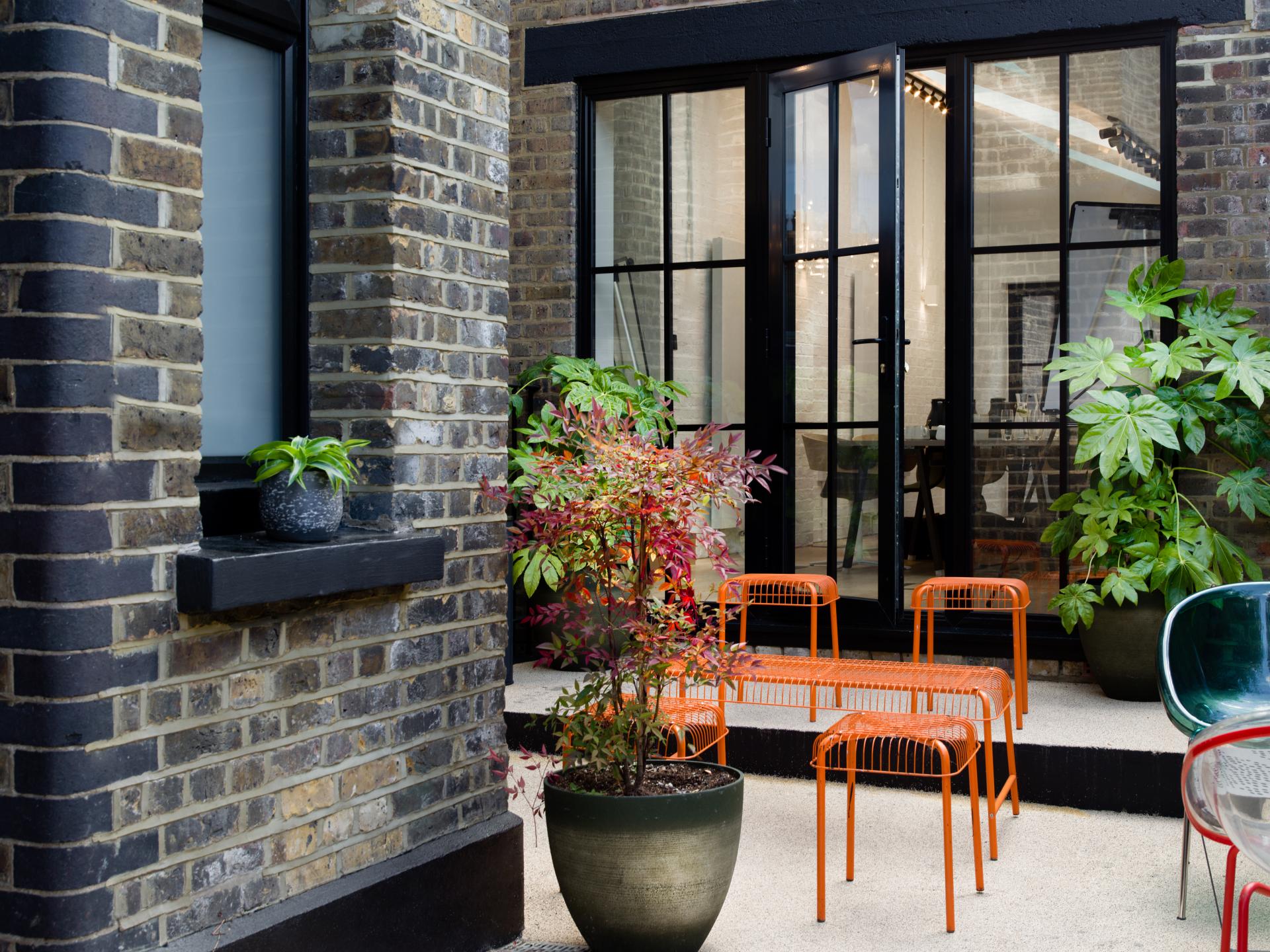 Stylish outdoor workspace featuring vibrant orange chairs, potted plants, and modern glass doors for flexible workplace design.