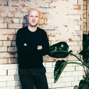 Matt Welsh, co-founder of TextileMania, standing in front of a brick wall with green plants, highlighting comfort and design.