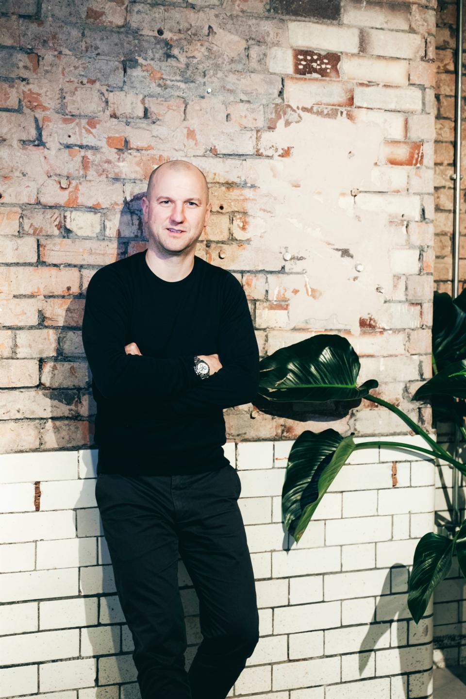 Matt Welsh, co-founder of TextileMania, standing in front of a brick wall with green plants, highlighting comfort and design.