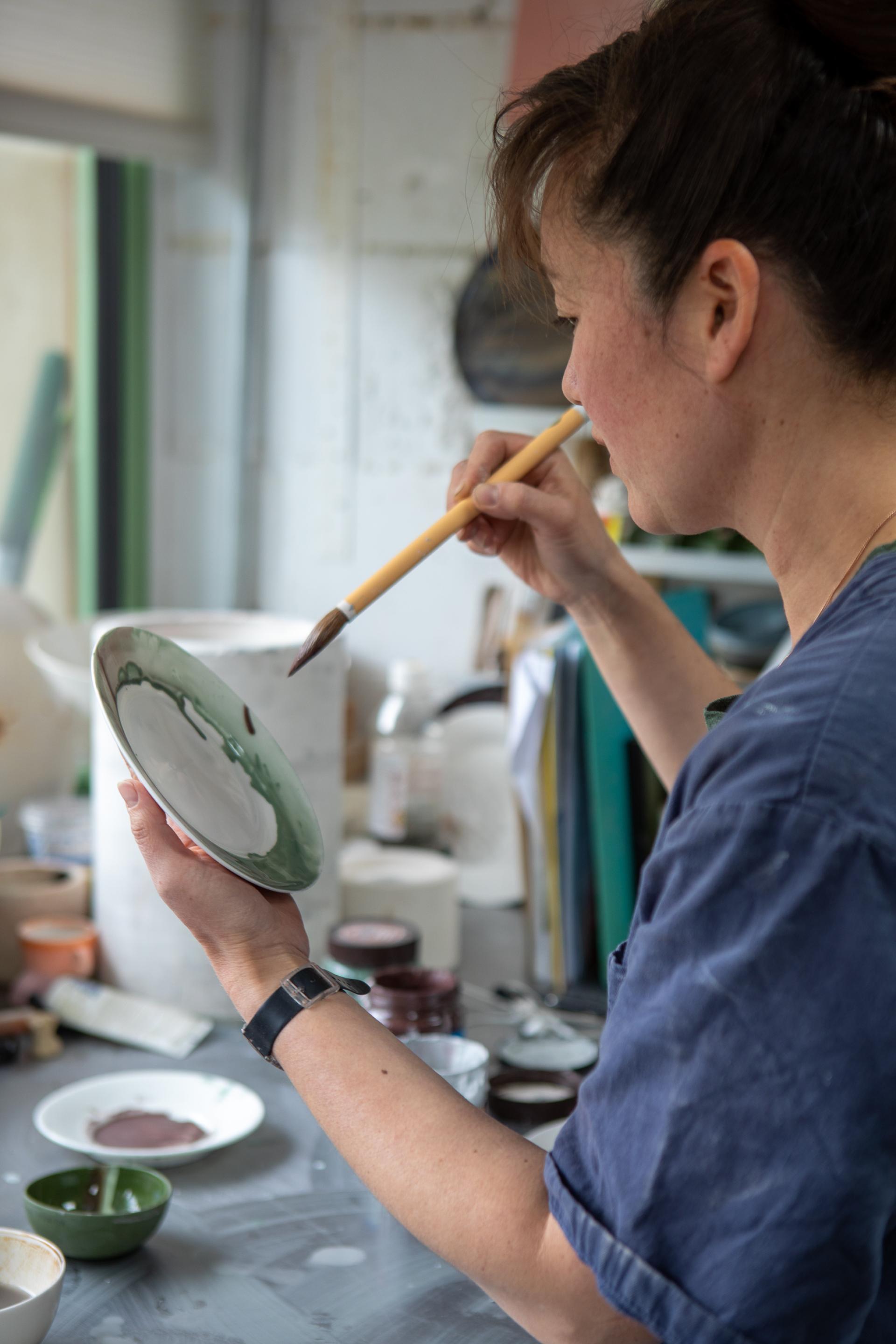 Reiko Kaneko skillfully paints a ceramic plate in her artistic studio, surrounded by various pottery materials.