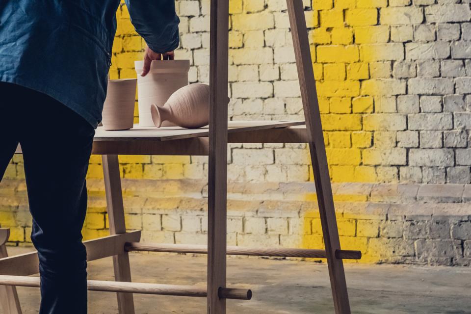 A person arranging minimalist pottery on a flexible wooden furniture display against a vibrant brick wall backdrop.