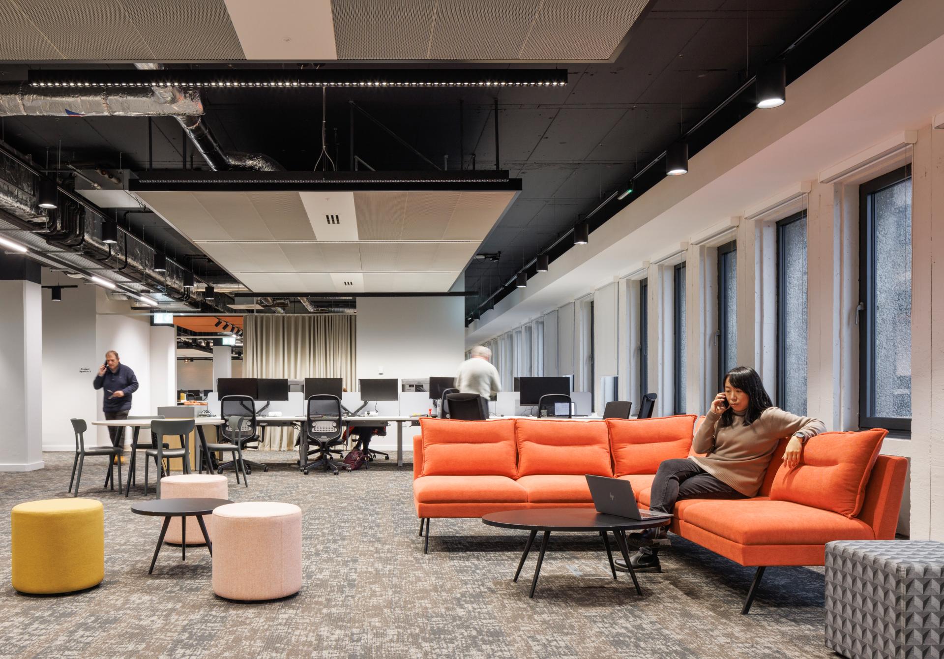 Modern office interior featuring vibrant orange seating, minimalist decor, and employees engaged in work at desks.