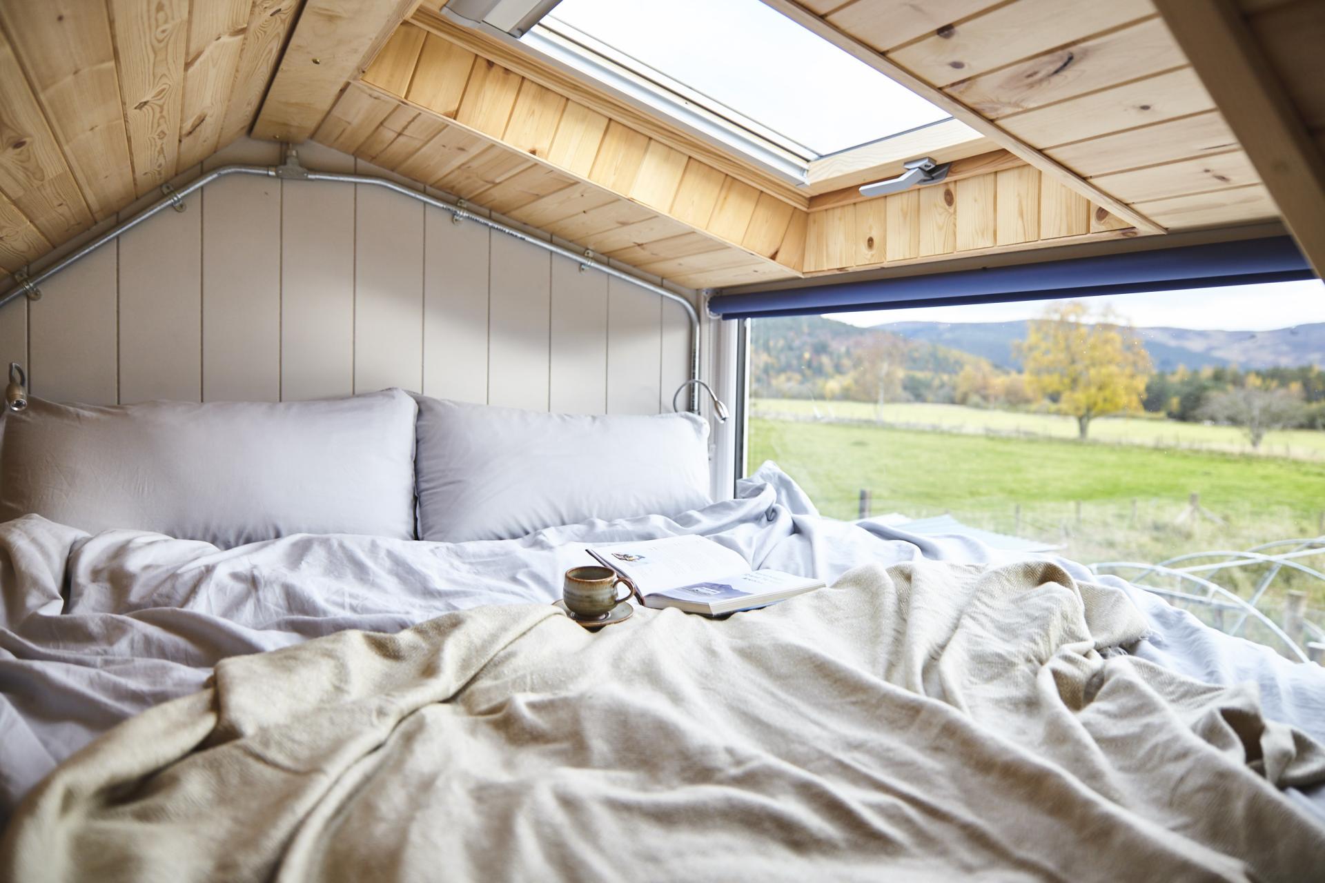 Cozy interior of a sustainable design space with a bed, blanket, book, and scenic view of nature.