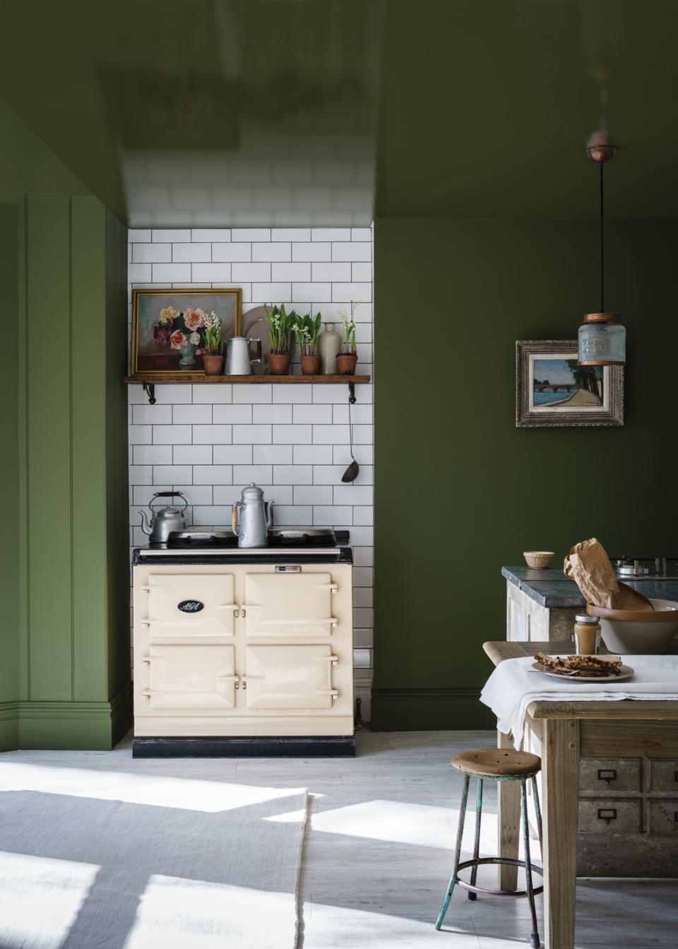 A stylish kitchen featuring a cream Aga cooker, green walls, and a rustic wooden shelf with plants and artwork.