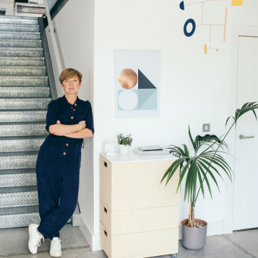 Kirsty Thomas stands in her design studio, showcasing Modernist decor, including geometric art and a minimalist furniture piece.