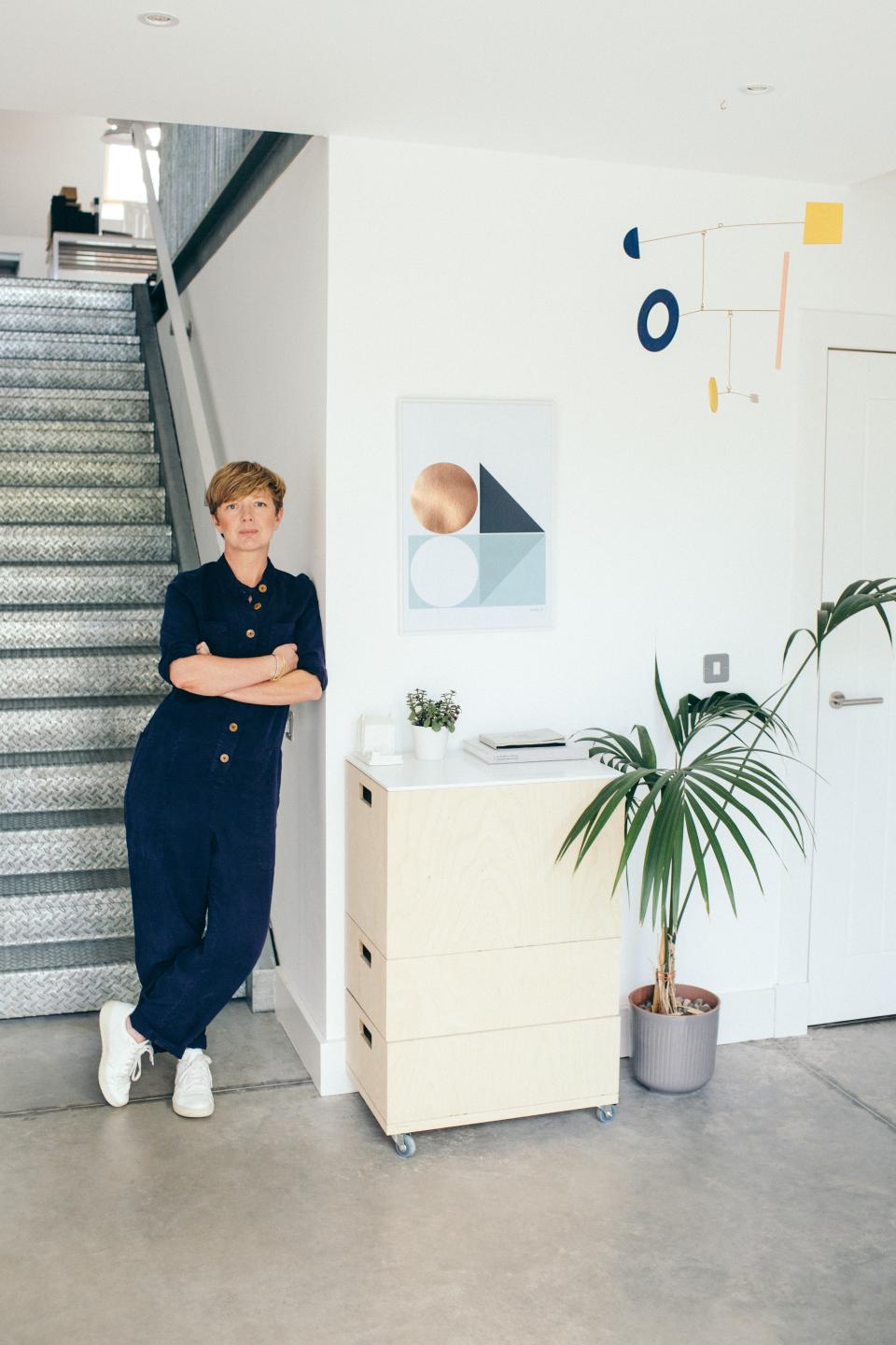 Kirsty Thomas stands in her design studio, showcasing Modernist decor, including geometric art and a minimalist furniture piece.