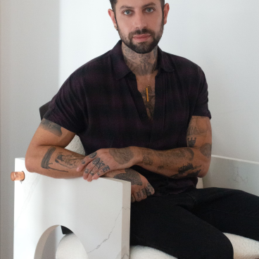 Evan Ward seated in a modern chair, showcasing his tattoos and distinctive style in a minimalistic setting.