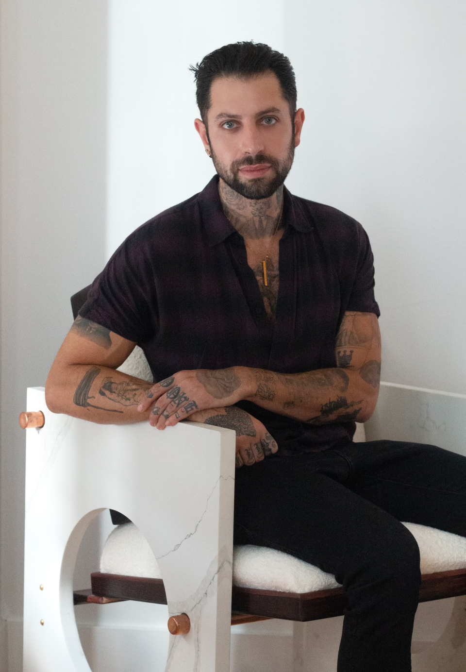 Evan Ward seated in a modern chair, showcasing his tattoos and distinctive style in a minimalistic setting.