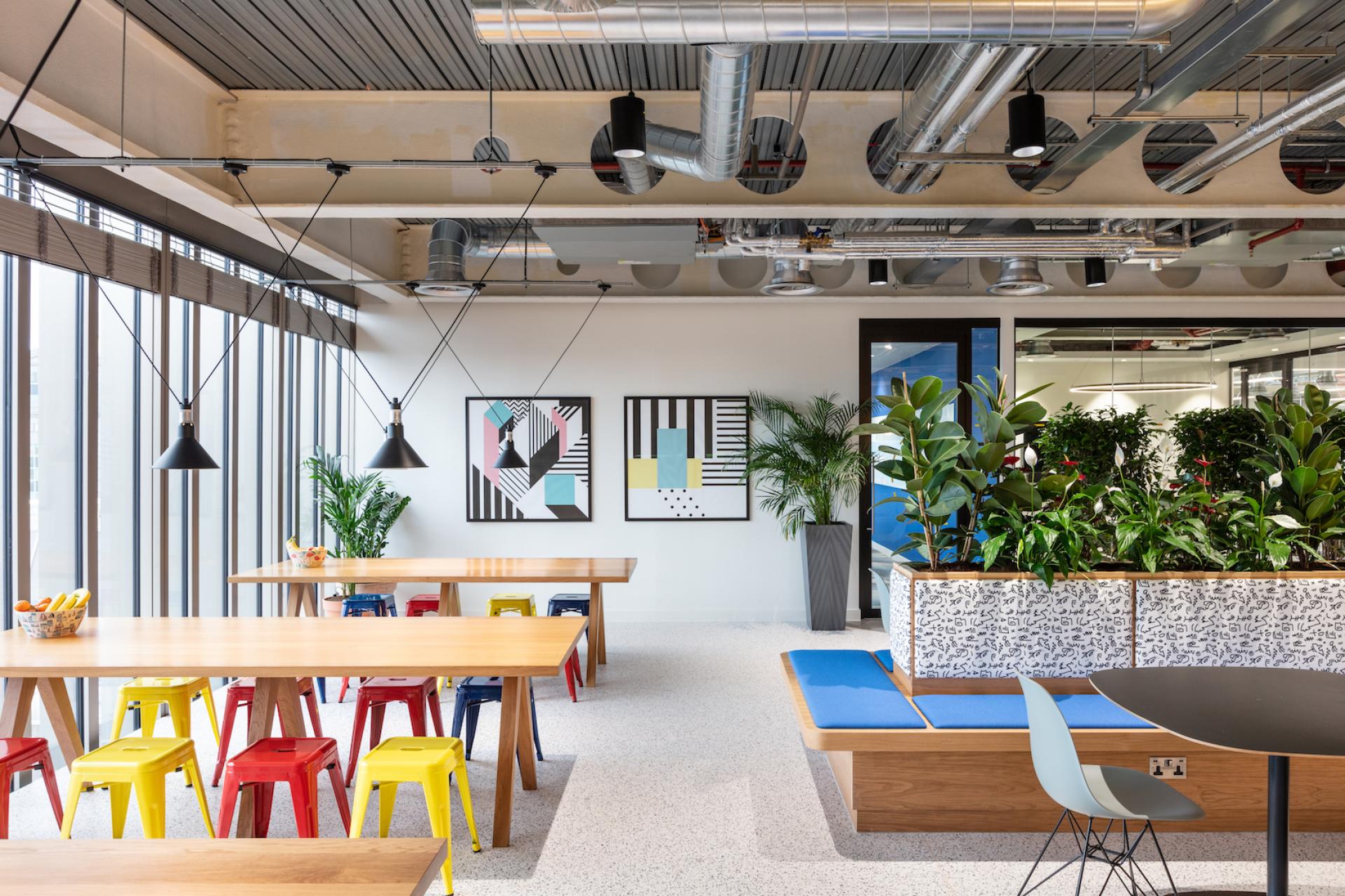 Modern office break room with colorful seating, hanging lights, plants, and contemporary artwork by Modus in Travelfusion's HQ.