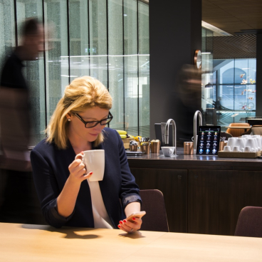 A woman enjoys coffee while using her phone in a modern office, highlighting Scanomat's premium TopBrewer partnership with QBE.