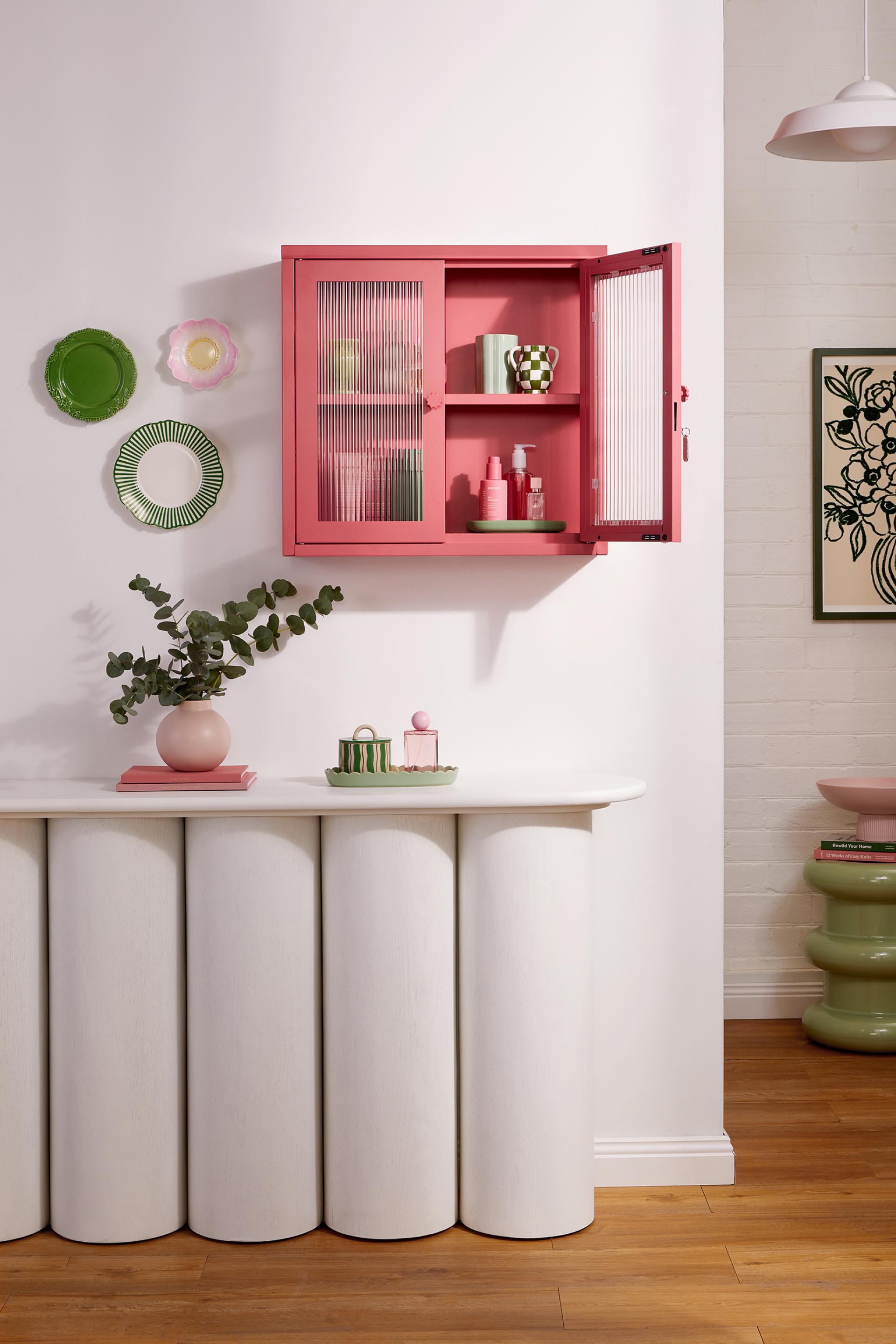 Colorful pink cabinet with open doors displaying decor items, accompanied by green plates and stylish tabletop accessories.