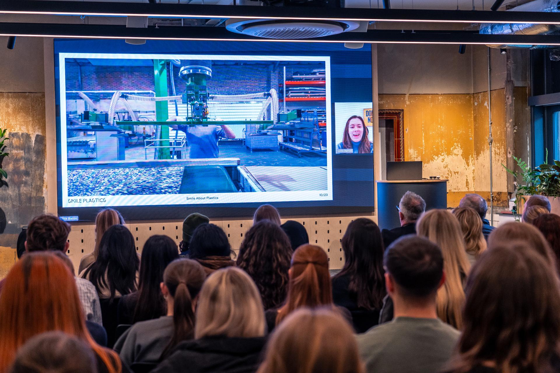 Audience attending a virtual presentation on sustainable design, featuring a factory image on screen about recycled plastics.
