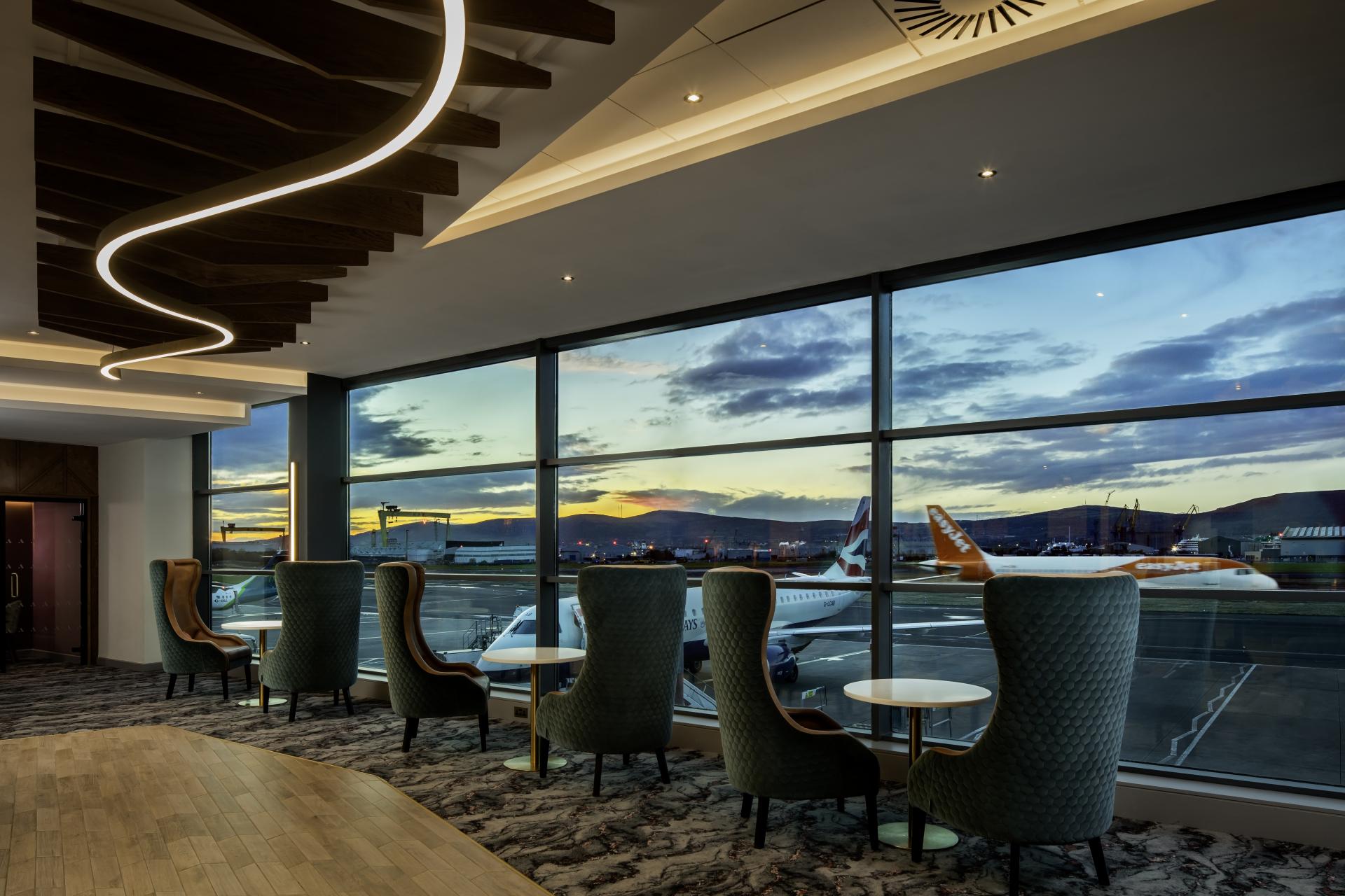 Modern aviation lounge with plush seating, overlooking Manchester Airport runway during sunset, highlighting design aesthetics and airport operations.