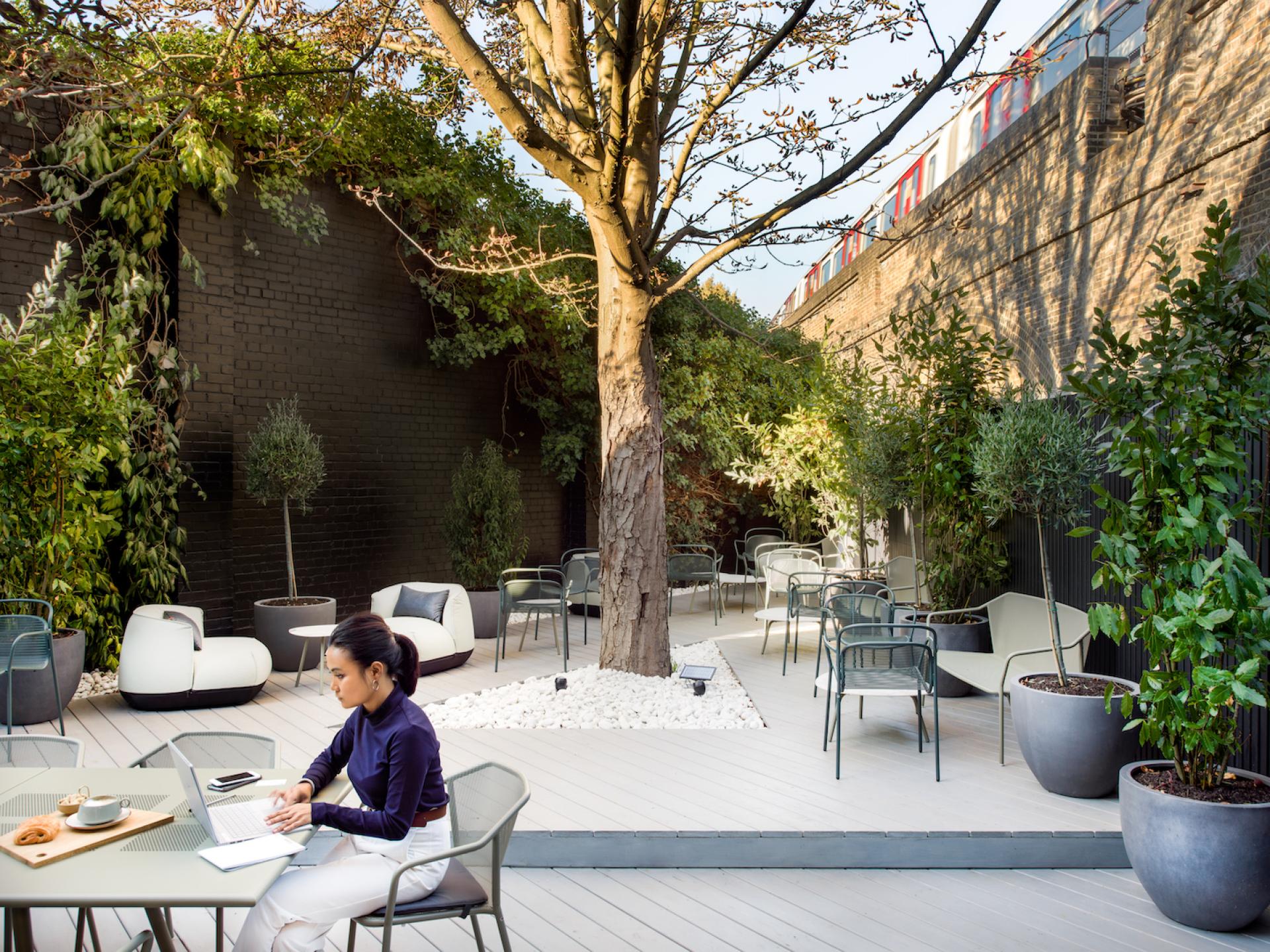 Urban outdoor workspace featuring a woman using a laptop, surrounded by greenery and modern seating for flexible work design.