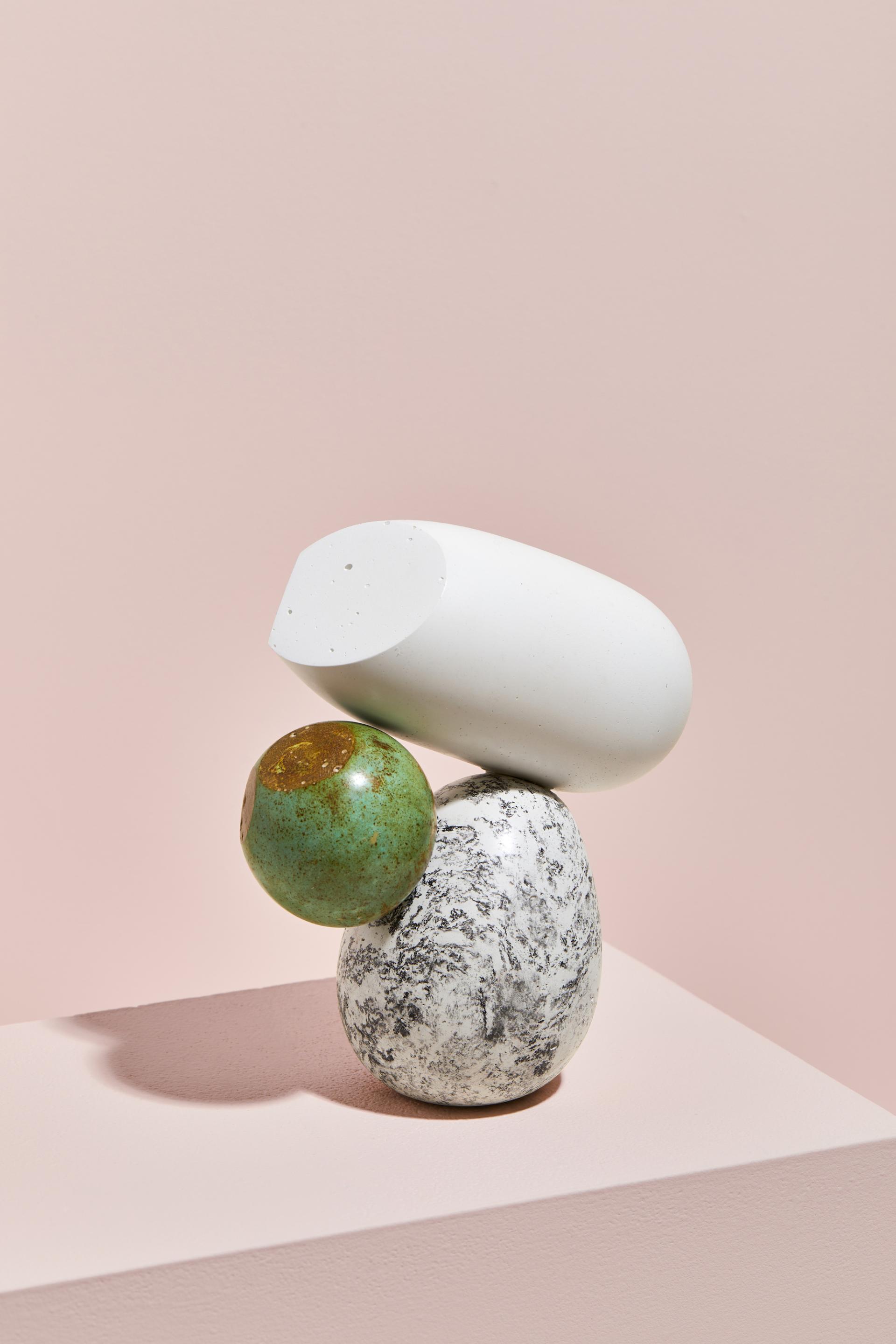 Abstract sculptural arrangement featuring a white shape, a speckled grey rock, and a green ball on a pink background.