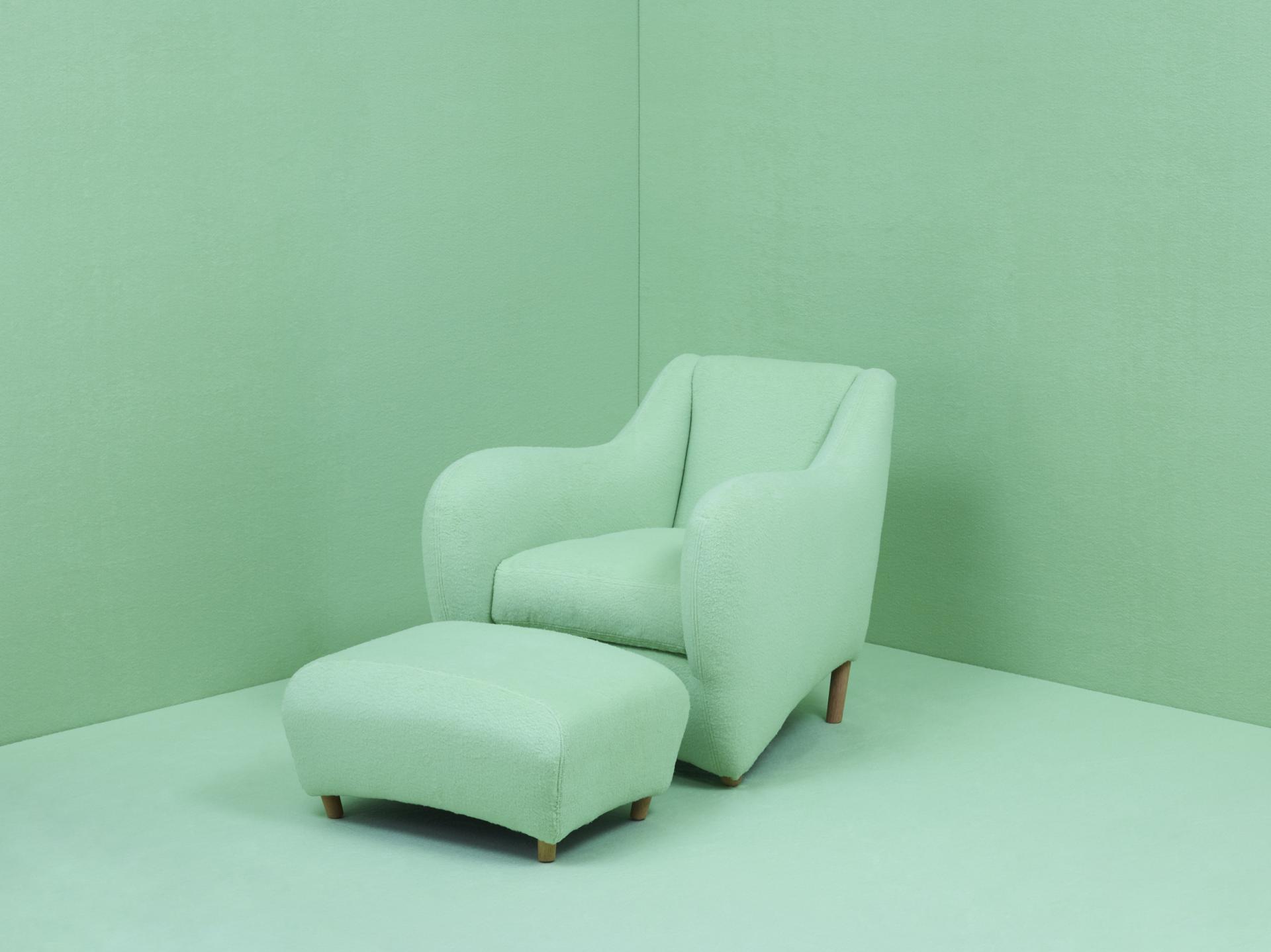 Matthew Hilton’s Balzac armchair with matching ottoman, celebrating thirty years of innovative design, against a soft green backdrop.