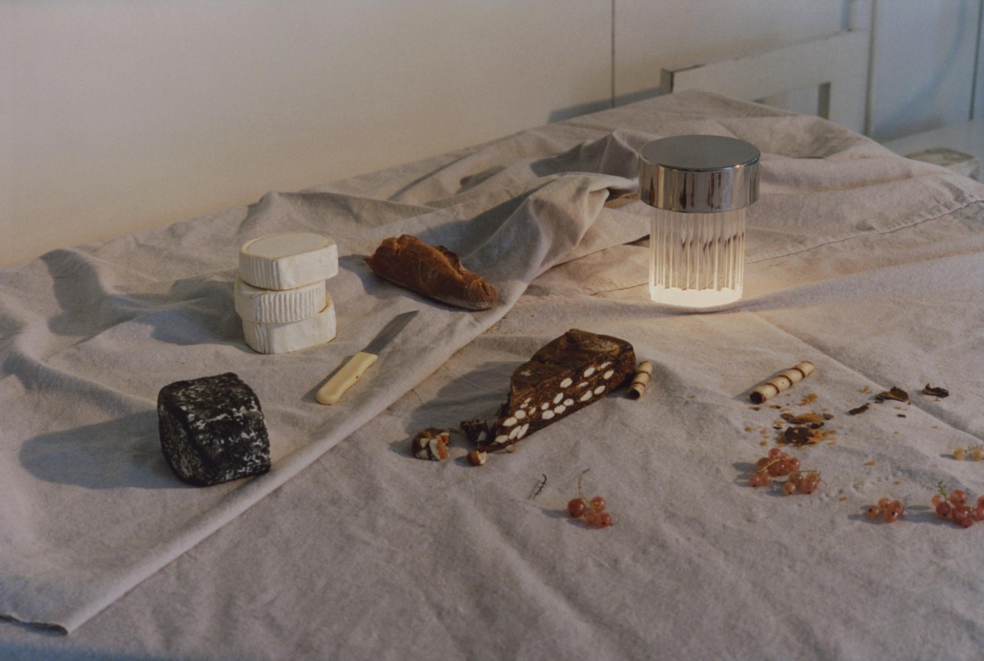 A stylish portable light illuminates a rustic table setting with cheeses, bread, and decorative snacks for outdoor dining.