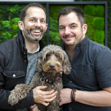 Two men holding a brown dog, standing in front of green moss wall art, showcasing authenticity and sustainability.