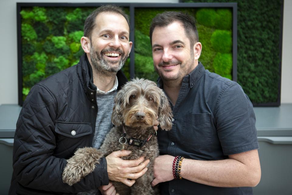 Two men holding a brown dog, standing in front of green moss wall art, showcasing authenticity and sustainability.