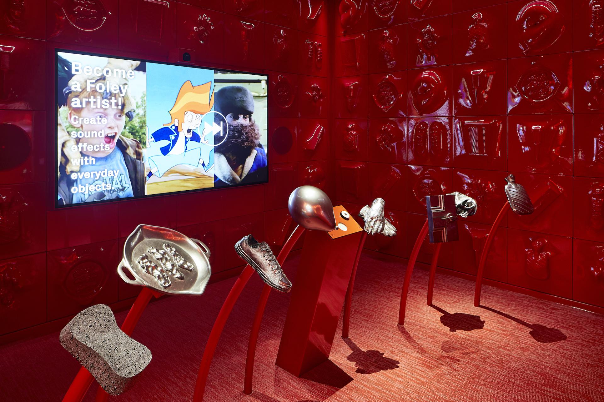 Interactive exhibit at ACMI features everyday objects for creating sound effects in film, surrounded by vibrant red walls.