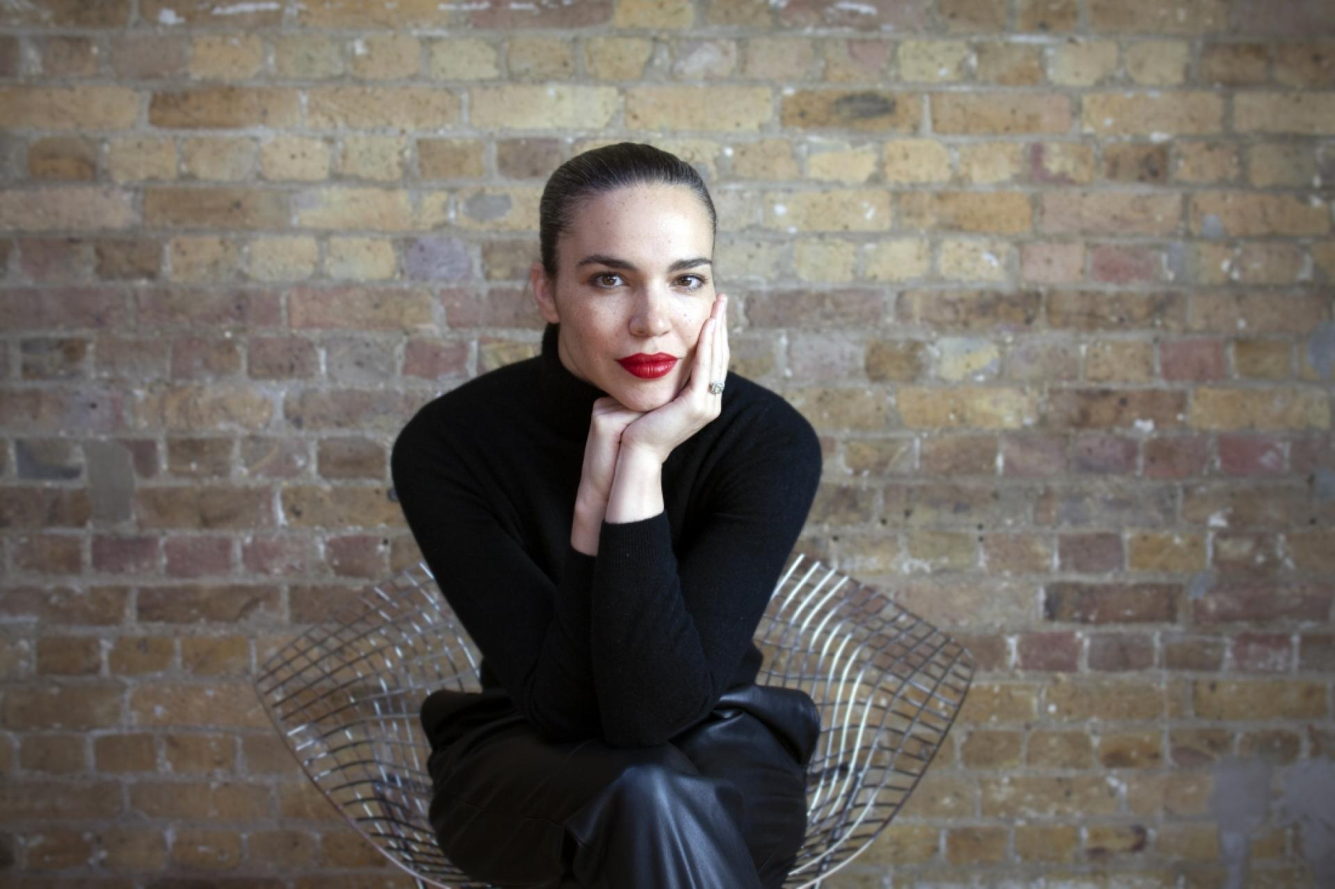 Sophisticated woman in a black turtleneck, seated in a modern chair against a brick wall, exuding confidence and inspiration.