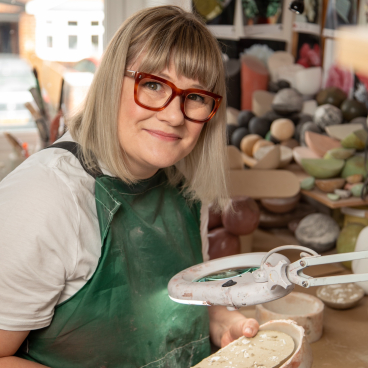 Sculptor Vic Wright in her studio, holding a piece of clay while surrounded by various sculpting materials.