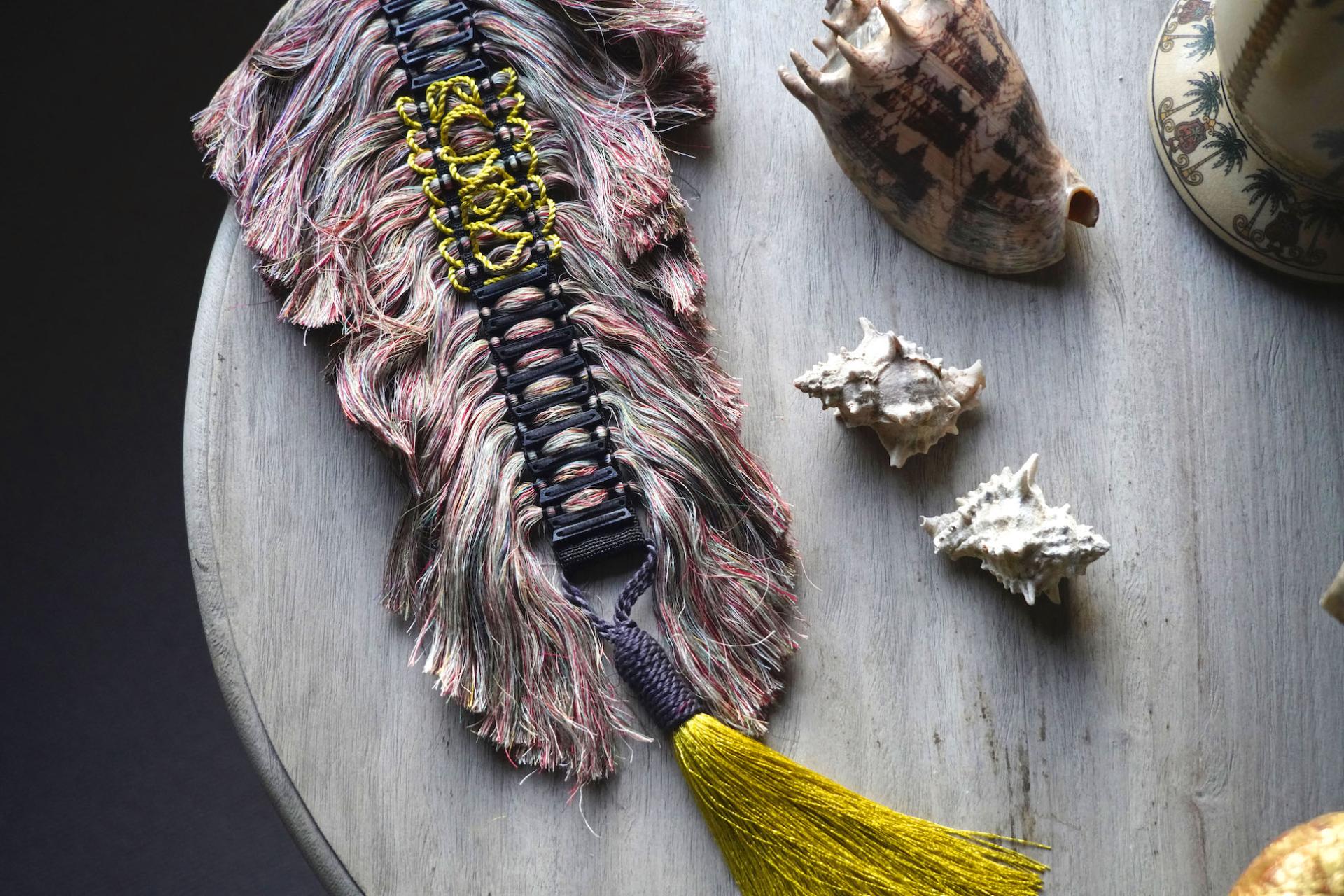 Colorful handmade textile artwork displayed on a wooden table, surrounded by decorative shells and artistic objects.