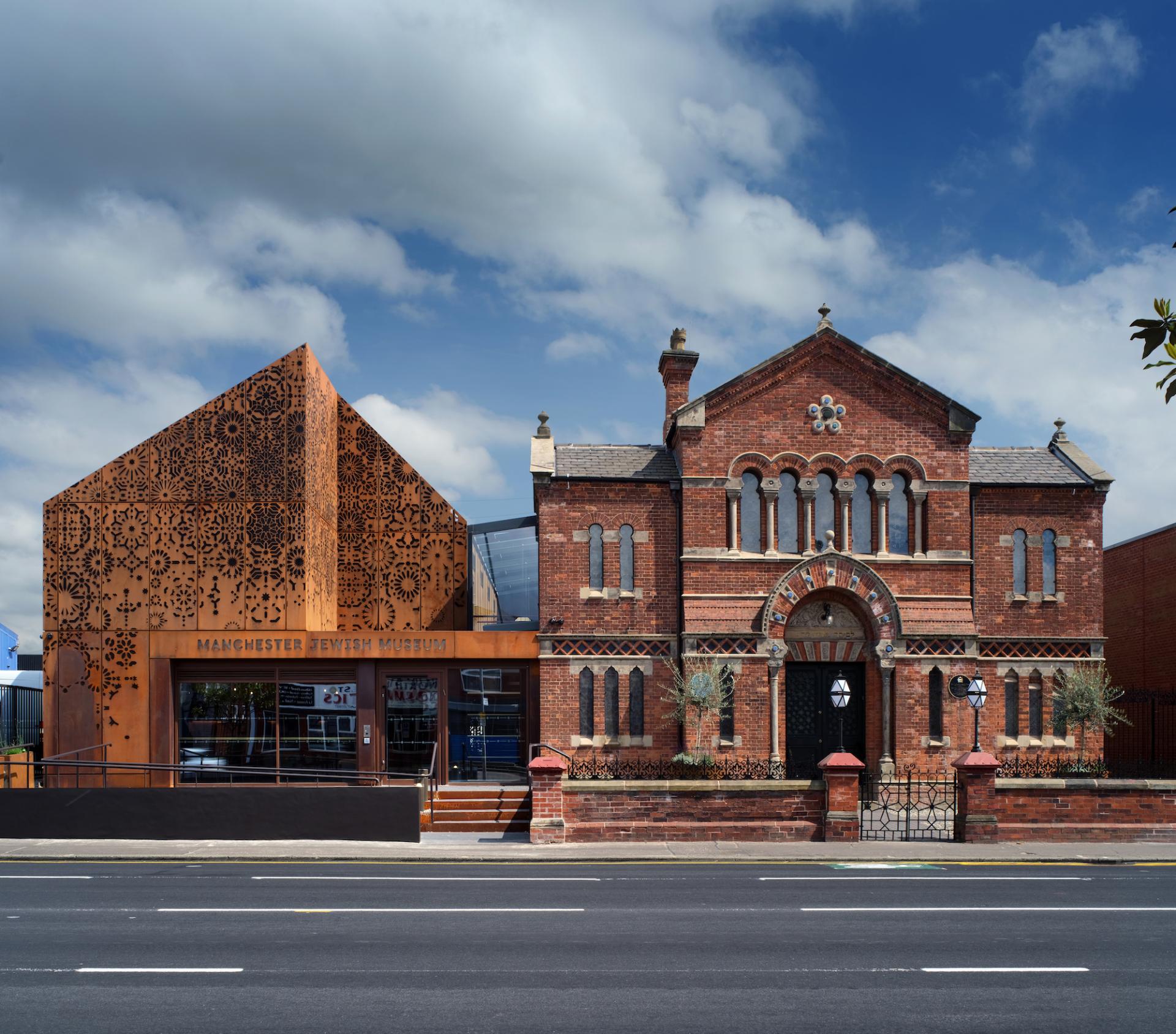 Modern extension of Manchester Jewish Museum features corten steel facade and historic red brick architecture against a vibrant sky.