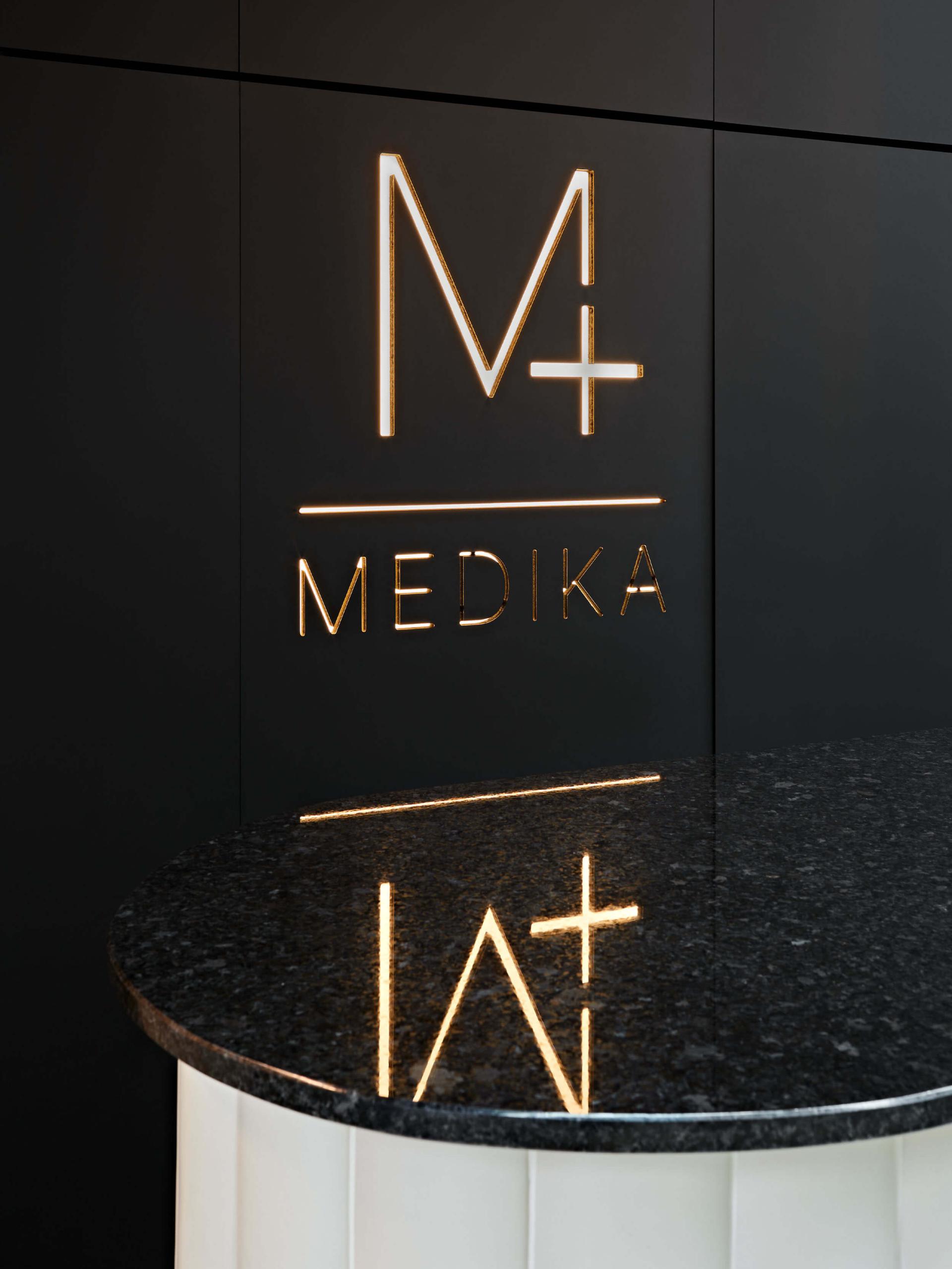 Medika Clinic's stylish reception area featuring the illuminated logo against a sleek black wall and polished stone counter.