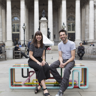 Two designer-makers seated on a colorful modern bench in an urban setting, highlighting contemporary design and craftsmanship.
