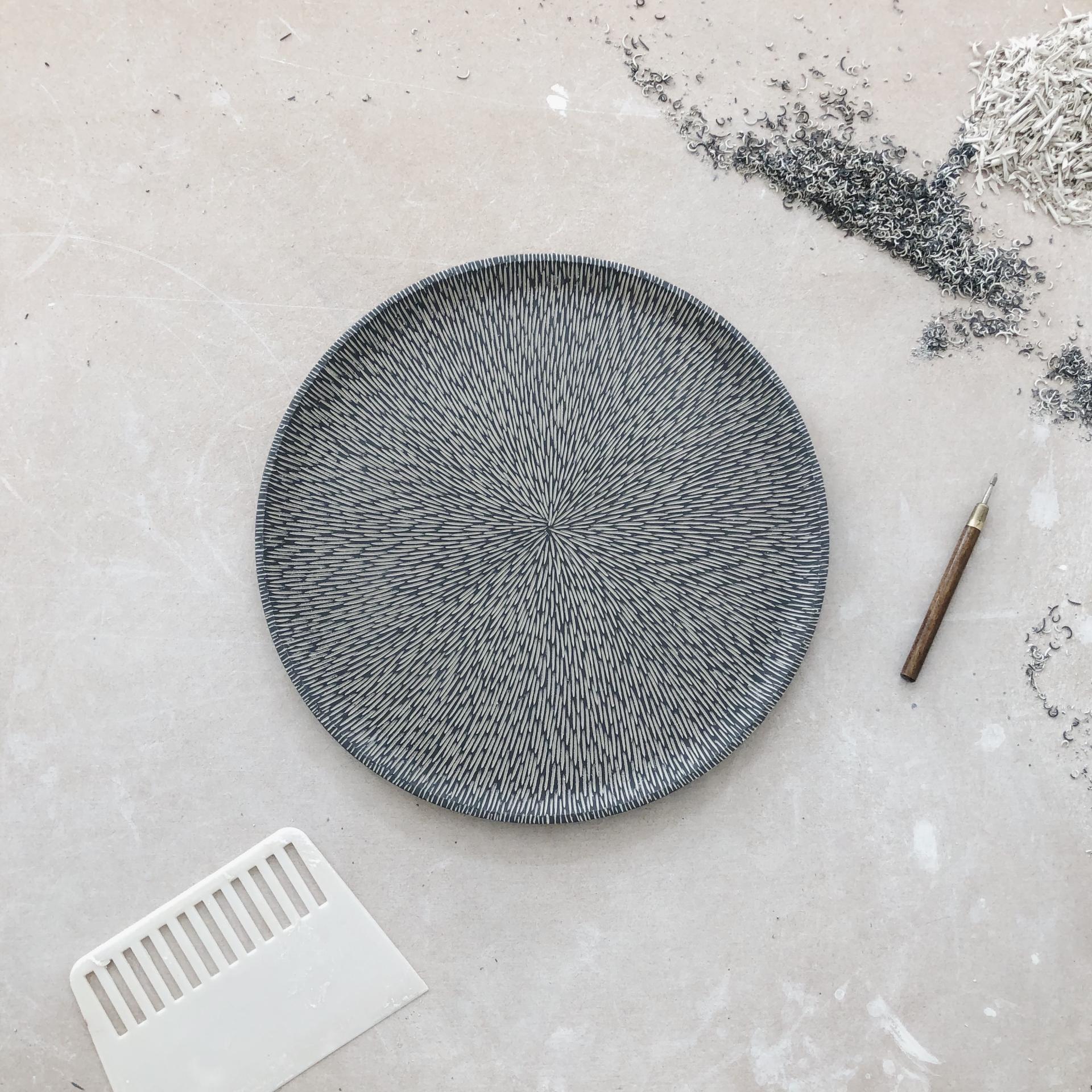 Handcrafted ceramic plate with intricate line patterns, surrounded by sculpting tools, showcasing Sophie Farrar's artistry in pottery.