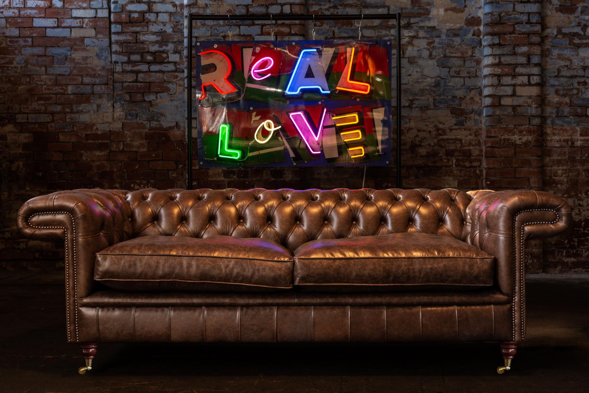 Leather Chesterfield sofa in an industrial setting, featuring a vibrant "REAL LOVE" neon sign backdrop.
