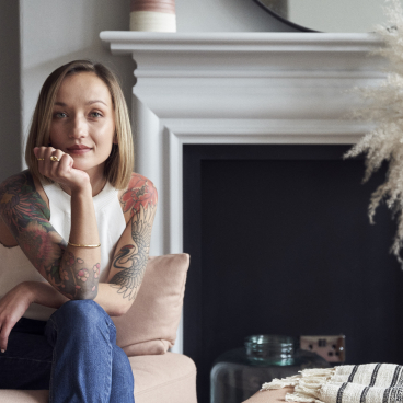 Chloé Cotard sits thoughtfully on a sofa, showcasing her vibrant tattoos in a stylish interior design setting.