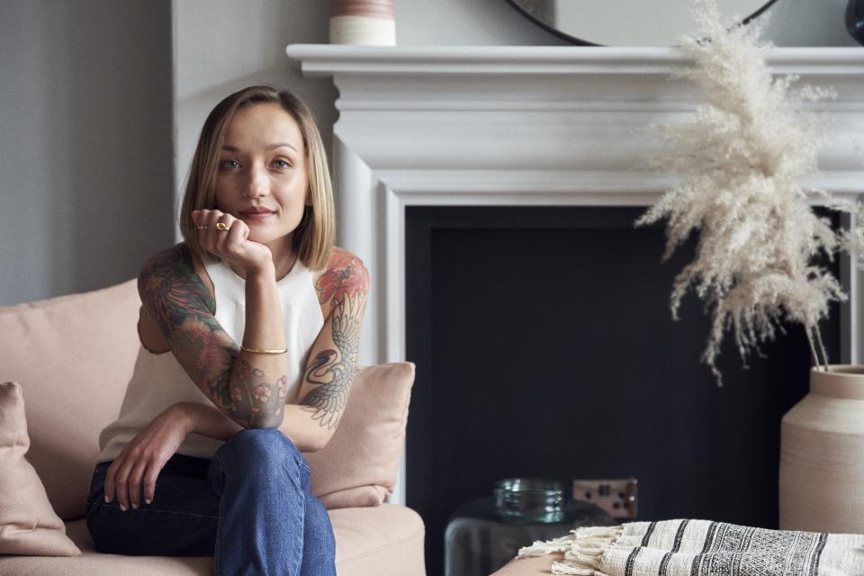 Chloé Cotard sits thoughtfully on a sofa, showcasing her vibrant tattoos in a stylish interior design setting.