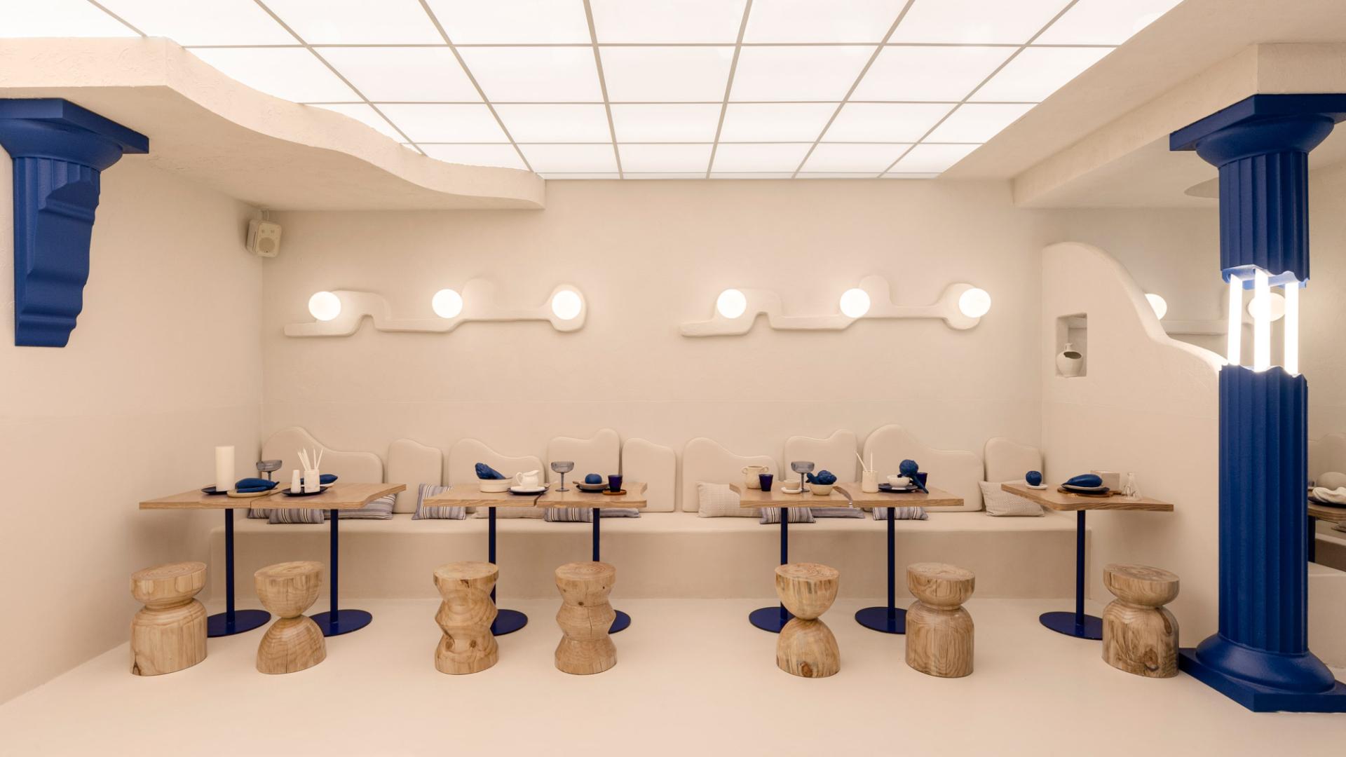 Contemporary Greek restaurant interior featuring minimalist design, wooden stools, and blue accents in a bright, inviting space.