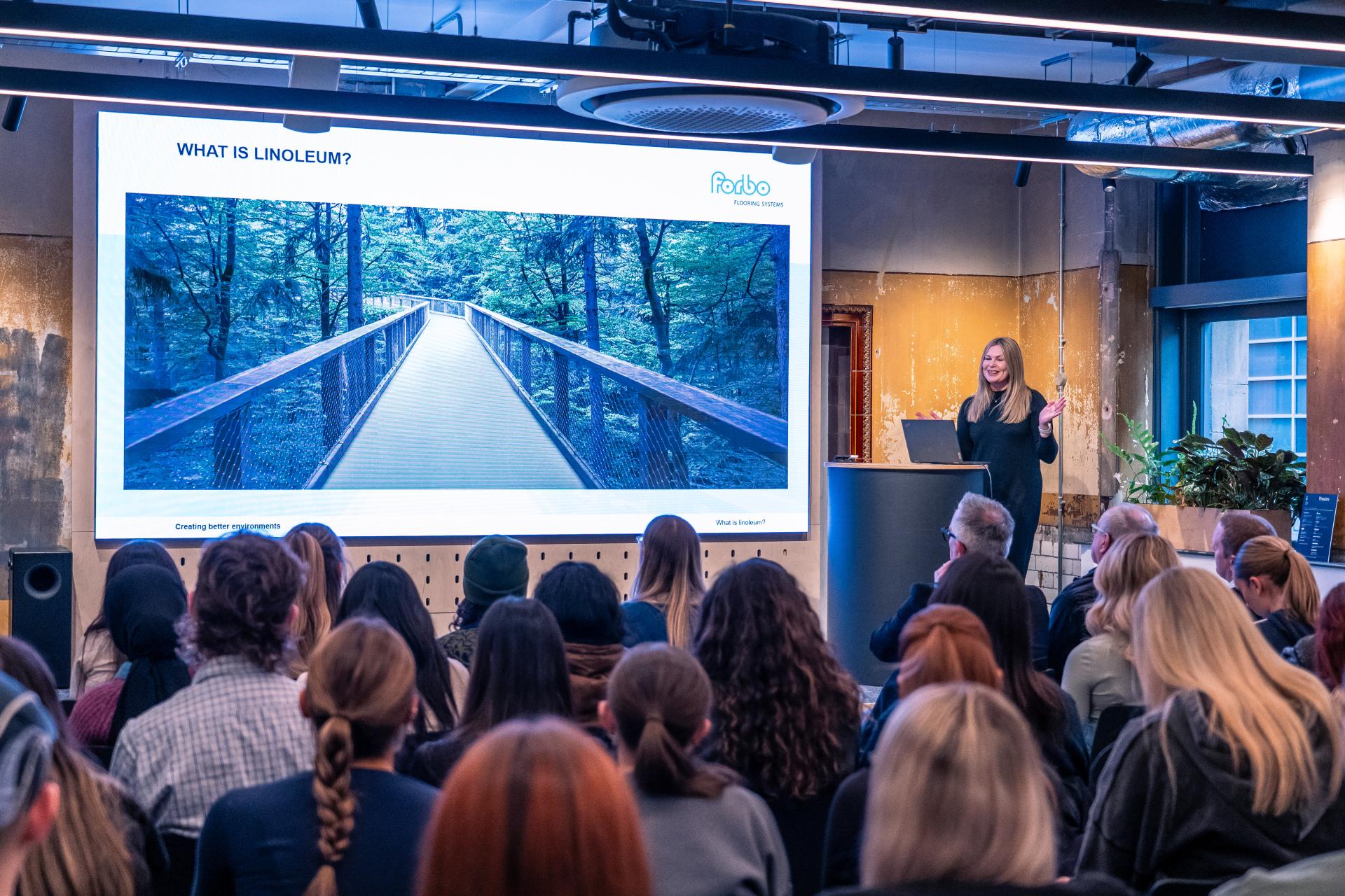 Interior design presentation with a speaker discussing linoleum, showcasing a timber bridge in a forest setting.