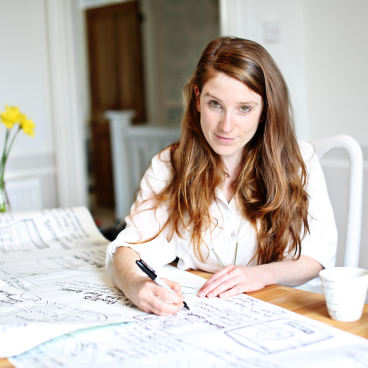Charlotte Cosby sketches design ideas on a table, surrounded by flowers and a refreshing drink, embodying creative inspiration.