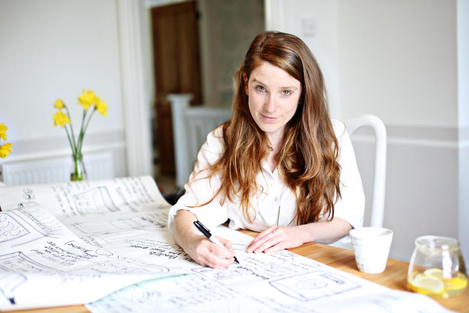 Charlotte Cosby sketches design ideas on a table, surrounded by flowers and a refreshing drink, embodying creative inspiration.