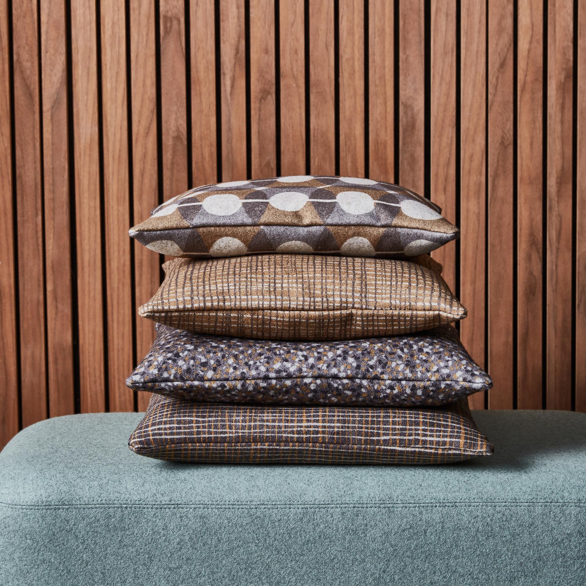 Textured wool cushions in various patterns stacked against a wooden backdrop, showcasing Camira's innovative printed wool collection.