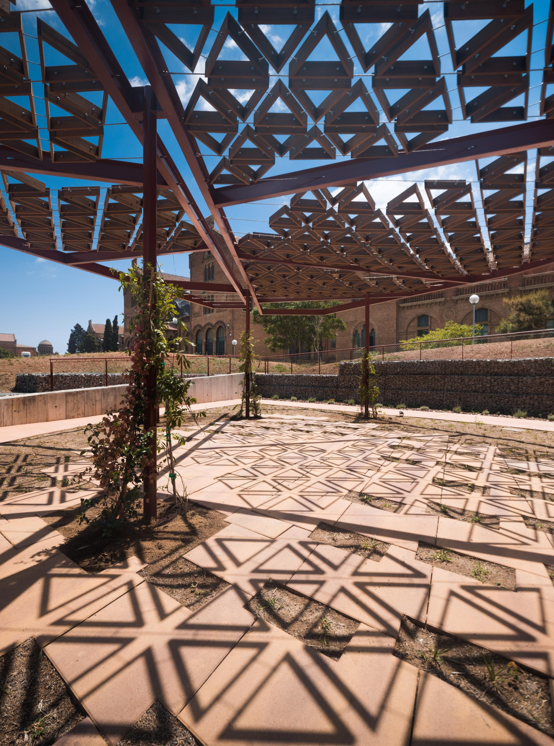 Thermally modified tulipwood canopy creates intricate shadows over the patterned ground at Kālida Sant Pau, Barcelona.