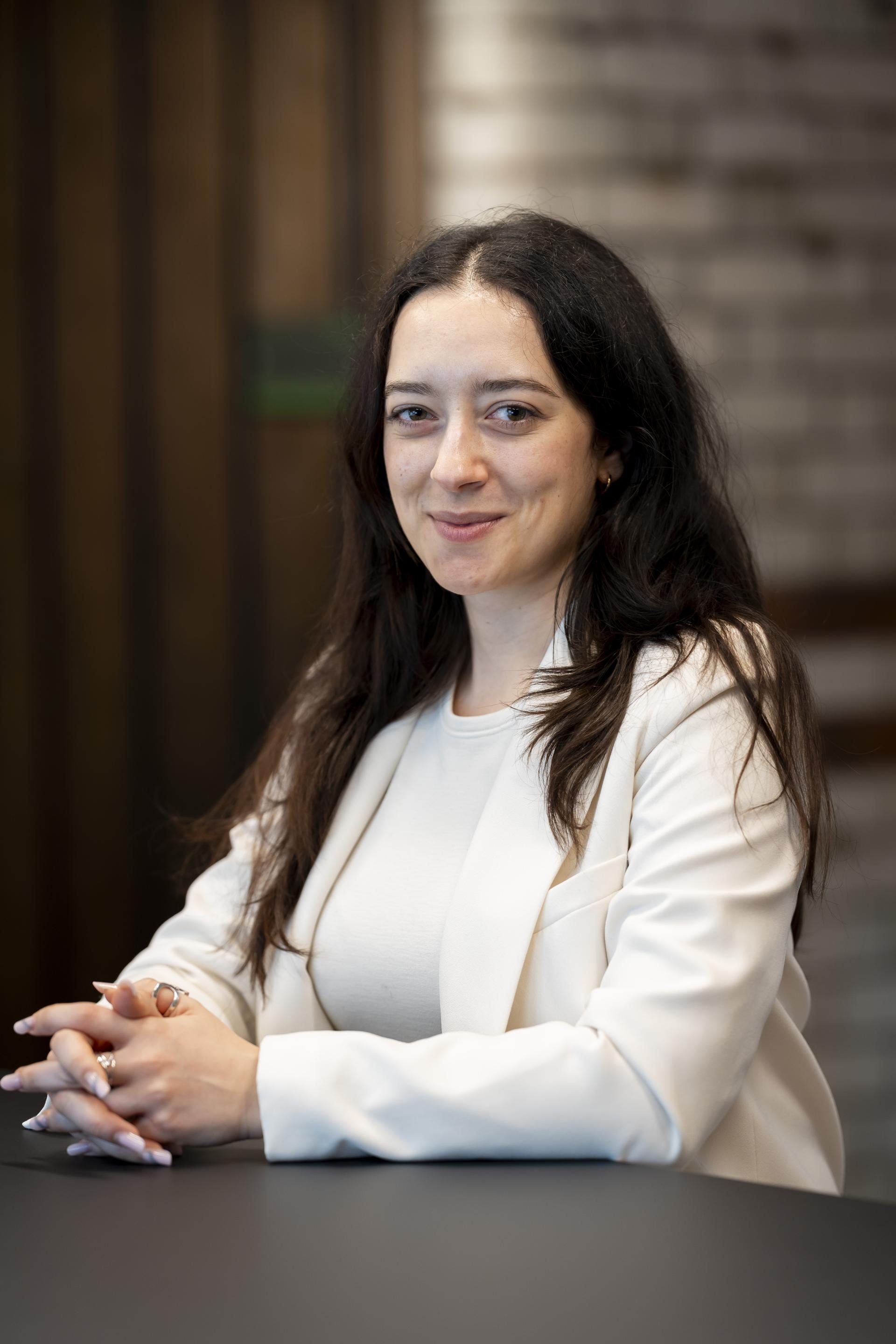 A professional woman in a white blazer sits at a table, smiling, discussing technology's role in sustainable design.