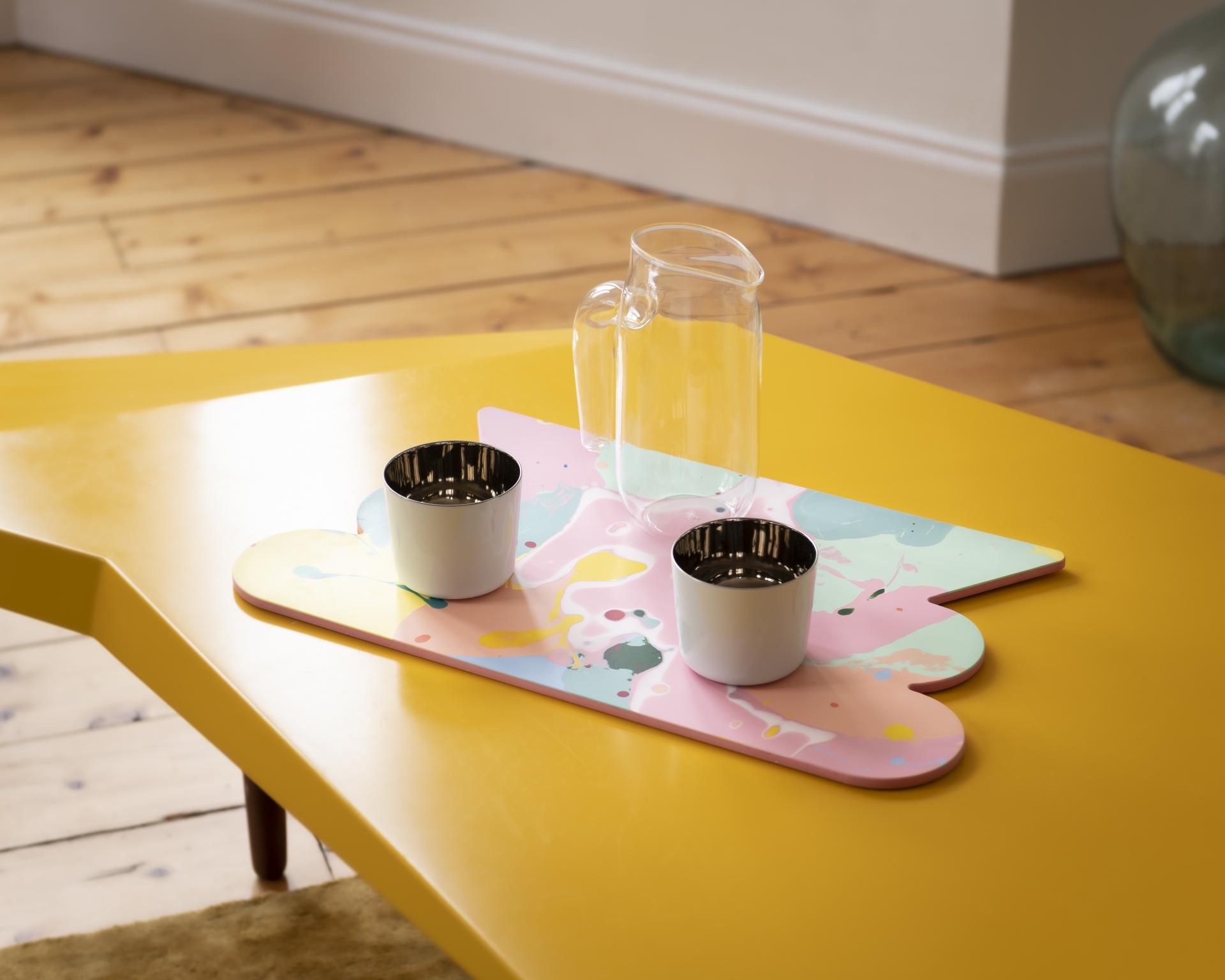 Vibrant surface design featuring a pink tray, playful tableware, and a glass jug on a yellow table.