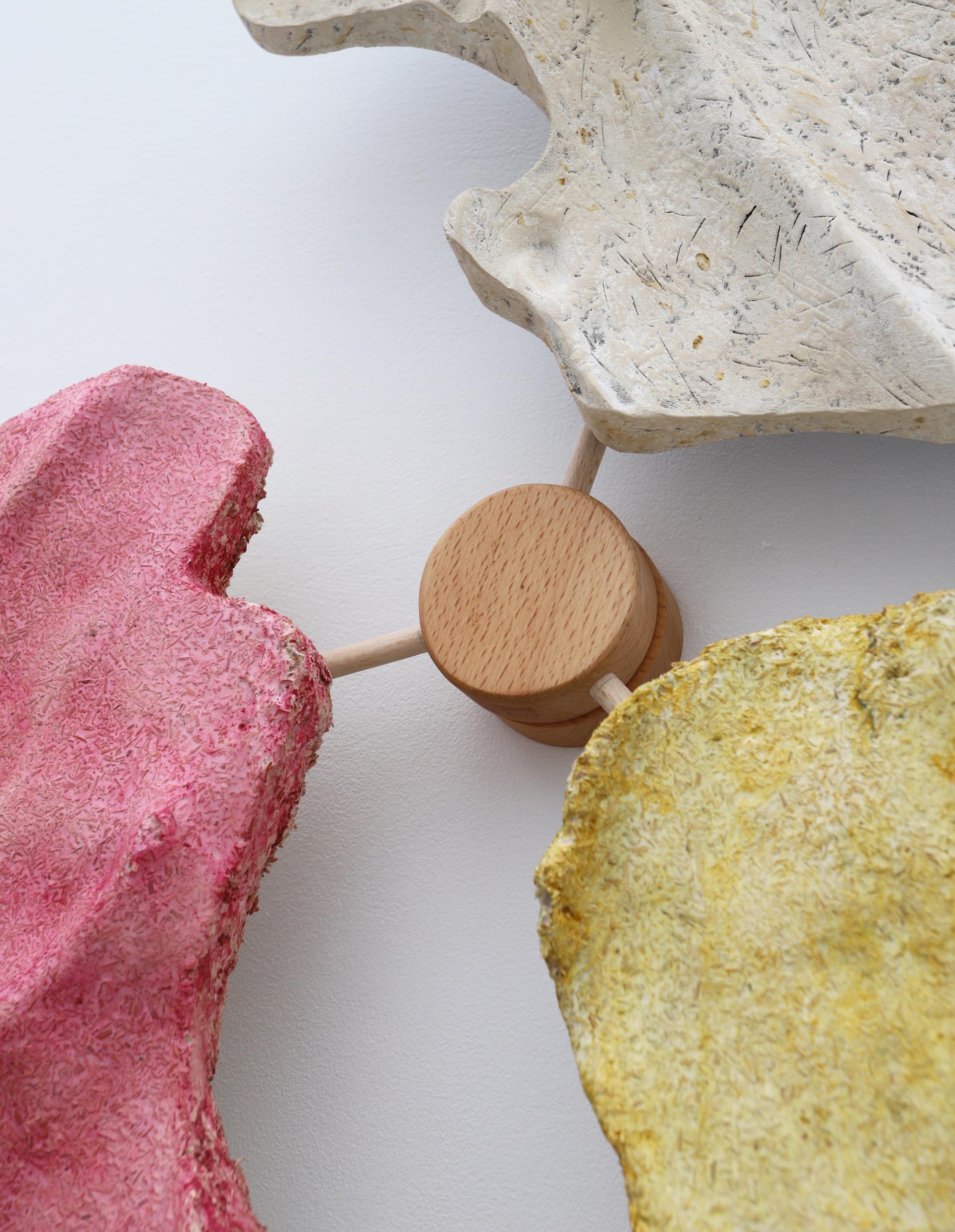 Colorful textured art pieces in pink, yellow, and white arranged around a wooden central support for New Designers 2024 exhibition.