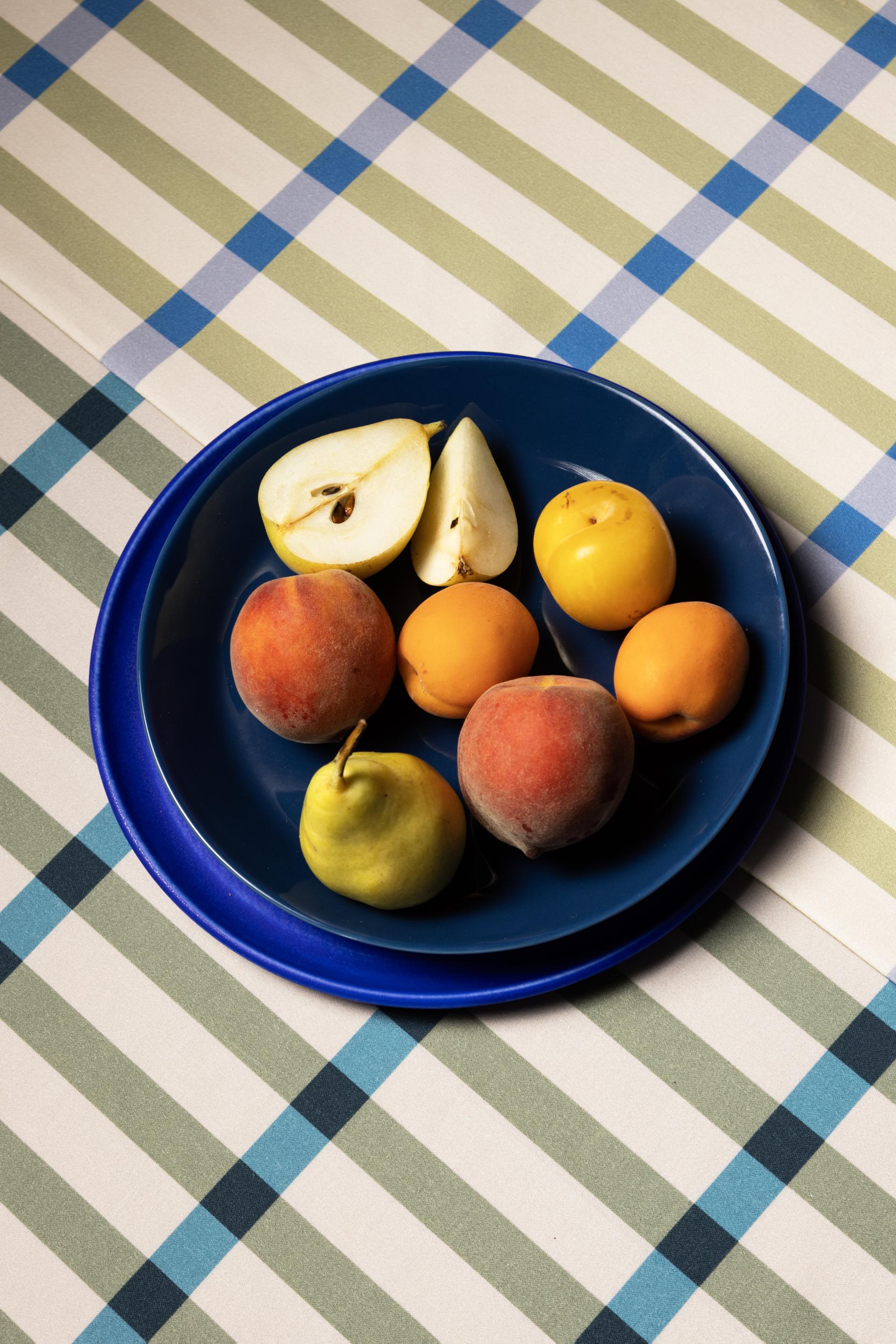 Colorful fruits including peaches, pears, and apricots arranged on a blue plate atop a patterned tablecloth.